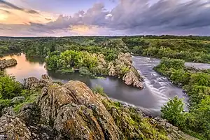 A photograph of a river through the forest at sunset, with orange lichen-covered rocks in the foreground, a purple and yellowish pink sunset sky, a river reflecting the sky colors, and bright green trees and plants.