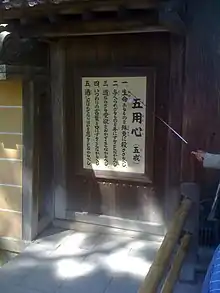 Plaque with Chinese characters written down vertically. A male hand is pointing toward them with a pointer.