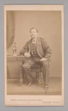 Frontal portrait of a seated Victorian man with his right arm leaning on a table and holding a pen