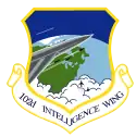 A standard shield, surrounded on the edge by a diminished yellow border. The upper part of the shield is blue, with one gray aircraft with a white trail pointing out from the left side. The plane is flying over the northeastern portion of the western hemisphere, which is colored in green and light blue. On the front of the gray aircraft with a white outline is a cockpit, with the jet firing three rockets. The three rockets trail white smoke and are aimed the same direction as the aircraft. Under the nose of the plane are two black aircraft coming out of clouds and rising up towards the jet. Attached to the shield is a white scroll edged with the same color border as the shield, surrounding the inscribed "102D INTELLIGENCE WING" in blue