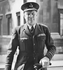 Half-portrait of man in dark military uniform with peaked cap, carrying gloves in left hand