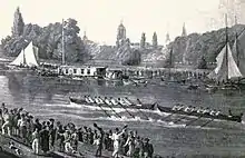 A river scene, with two eight-oared boats racing in the middle of the river, one just in front of the other. There are crowds on each bank and some sailing boats and barges by the far bank. In the distance, trees and church spires.