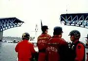 Rescue workers wearing orange uniforms on the boat approaching the bridge collapse. The two anchor trusses can be seen standing, while the middle span of the bridge is completely missing.