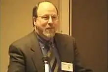 A middle-aged man with balding blond hair, a white beard, and glasses stands beside a microphone in his grey suit and pattern tie.