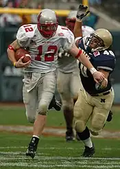 An American football player holding the ball in a white uniform with red lettering evades a tackle by another player in a blue uniform with white lettering.