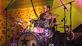 A colour photograph of Starr playing a dark coloured drum kit on a stage. The background is yellow.