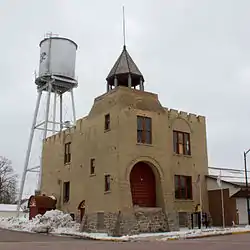 Two-story brick building with a buttressed stone foundation, crenellated parapet, and open belfry