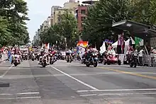 Dozens of motorcycles riding down a street, with rainbow flags visible in the background.