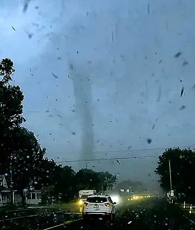 Central cone of tornado surrounded by flying debris on the horizon. Below horizon is a street with vehicles stopped. It is raining.