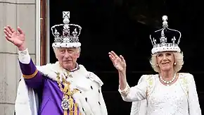 Charles and Camilla wearing their crowns and coronation robes waving from the balcony of Buckingham Palace