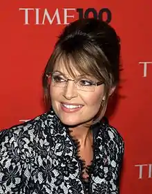 A woman with brown hair tied back, and glasses, looks to her side slightly, while speaking into a microphone