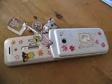 A Japanese flip phone typical of the 2000's with phone charms. These phones and charms were symbolic of youth culture in the early Heisei era, and remained prevalent in Japan long after the advent of the smartphone made flip phones obsolete elsewhere.