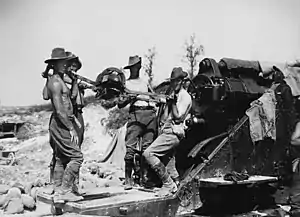 Bare-chested men in slouch hats and breeches stand by a large artillery piece, ready to load a shell.