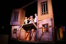 A woman performs a puppet show by manipulating puppets that are situated on a stage, stylized as a wooden playhouse.