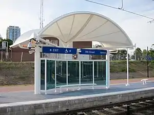 A train stop in North Carolina, with a white canopy, curved blue glass windscreen, and metal bench.