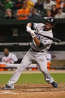 A man in a grey baseball uniform with "Rays" on the chest and "TB" on his blue batting helmet winces as he stops his swing.