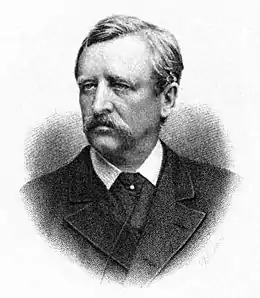 Head and shoulders portrait of a middle-aged man, facing half-left. He has dark, neatly brushed hair, a heavy moustache, and is wearing a dark, formal jacket.