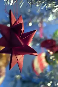 An out-of-focus close-up of a part of a Christmas tree with glitter garlands and blurry decoration. In-focus in the left half of the picture, suspended from one of the twigs, is a red cardboard-woven Froebel star. Four tips and seven prongs are visible.