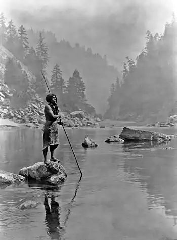 A smoky day at the Sugar Bowl—Hupa, c. 1923. Hupa man with spear, standing on rock midstream, in background, fog partially obscures trees on mountainsides.