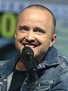 A young man with a buzzcut wearing a denim jacket and a black shirt faces the camera, speaks into a microphone.