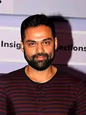 A middle-aged Indian man wearing a black-and-red-striped shirt smiling at the camera. He has short, black hair; a short, face-hugging beard and a moustache.