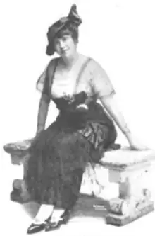 A white woman seated on a bench, wearing a dark hat and a dark long dress with a light underblouse; she appears to be corseted