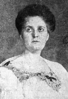 A white woman with dark hair in an updo, wearing a light-colored dress with a lace-embellished neckline