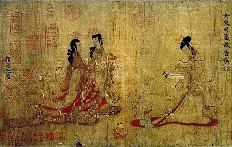 Two ladies walking towards another lady standing at a writing table with a writing brush in her hand