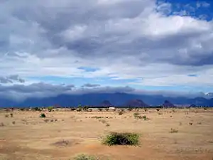 Daytime view of an almost lifeless expanse, dry rocks and sand marked only by the odd lone shrub. The dry terrain reaches to a chain of mountains in the far distance, near the horizon. A bank of clouds soars above the void, but it does not appear to hold the promise of rain. A far darker, larger, more turgid cloud bank sits above the and  distant mountains, above the horizon.