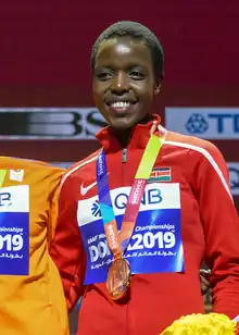 Agnes Tirop wearing a red athletic Nike jacket with a Kenyan flag, a 2019 World Athletics Championships placard, as well as her bronze medal, standing on a podium next to another contestant (not seen in photo), holding a bouquet of yellow flowers, grinning at audience.