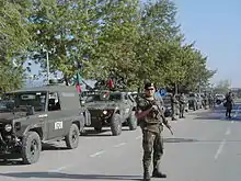 A soldier stand in front of a convoy of military vehicles, with KFOR markings and Portuguese flags. The second véhicule is a Portuguese VBL.