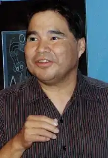 A middle-aged man with black hair wearing a collared shirt stands in front of a drawing on the wall.