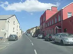 A road between two buildings, one red the other pebbledash, with cars parked on either side.