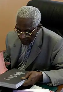 An image of Aimé Césaire in 2003 on a desk reading the cover of a book.