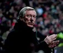 A side-on photograph of a man with grey hair. He is wearing glasses and a black overcoat.