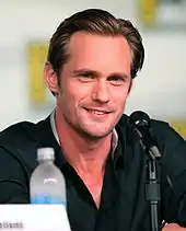 A man with blond hair wearing a darker-colored shirt smiles as he sits at a table