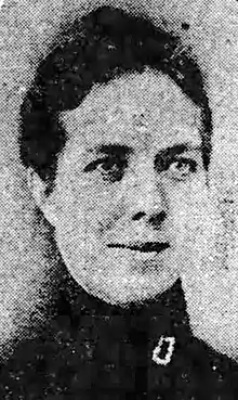 A white woman with dark hair dressed back, wearing a high-collared dark blouse or dress. She appears to have light eyes, and is slightly smiling.