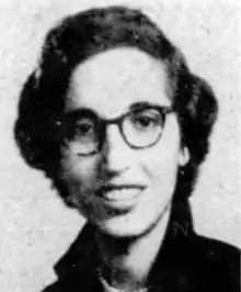 A young woman with dark wavy hair and olive skin, wearing dark glasses and a dark collared shirt or dress