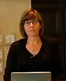 blurry image of White woman with shoulder-length hair, wearing dark outfit and glasses, looking at camera from behind laptop computer