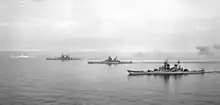 A black and white photograph of four large ships sailing on calm seas from the right to the left.