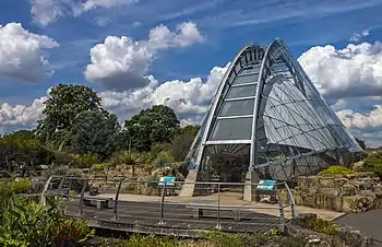 A narrow semicircular building of glass and steel latticework stands at the right, set amid an area of worked rock with a line of deciduous trees in the rear left, under a blue sky filled with large puffy white clouds. In front of it, curving slightly away to the left, is a wooden platform with benches on it and a thin metal guardrail in front of a low wet area with bright red flowers.