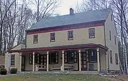 A two-story light yellow house with reddish trim, a pointed roof on the side, a small shed-roofed addition on the left and a porch along the front.