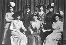 Seven white women (three seated, four standing) posed for a group photograph. They are all wearing long skirts and large hats. Several are wearing suit jackets.