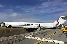 A jet airliner with tail-mounted engines and a T-tailed configuration, painted mostly white with a thin red stripe running along the fuselage below the windows, lying on its belly across a ditch; the aircraft's tail is resting on a paved road, while the forwards portion of the fuselage rests on a grassy field on the other side of the ditch.  The aircraft's tailcone has detached and is lying on the grass next to the left side of the fuselage.