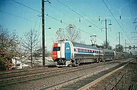 Stainless steel passenger rail cars with red and blue stripes horizontally across the windows. The visible end is covered with three vertical red, white, and blue stripes.