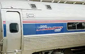 A stainless steel passenger rail car with a thick blue stripe across the windows and two thinner red stripes above. A logo within the blue stripe reads "Amtrak NortheastDirect Service".