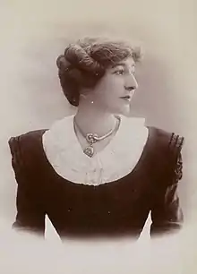 A white woman wearing a black dress with a white collar, and a chunky metallic necklace. Her shoulders are square to the camera but her face is almost in profile. Her dark hair is arranged back, away from her face and neck but covering her ears.