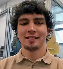 A young Caucasian man wearing a light brown collared shirt with dark brown curly hair and a slight beard on the chin