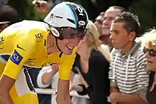 A road racing cyclist in a yellow skinsuit, wearing an aerodynamic helmet. His bicycle is not visible. Spectators watch from the roadside.