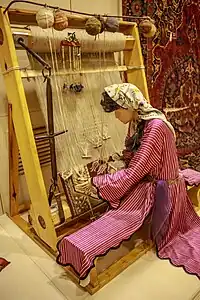 A Turkish carpet loom showing warp threads wrapped around the warp beam, above, and the fell being wrapped onto the cloth beam below.